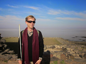 Picture of Justin, young blond man smiling and wearing sunglasses and a scarf and holding his white can at his side standing in front of a rocky beach with the coastline in the background
