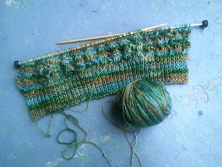 Flat Knitted lace on two wooden needles, with a bottom edge of ribbing. The yarn is a fingering weight, and varigated blue-green-brown. 