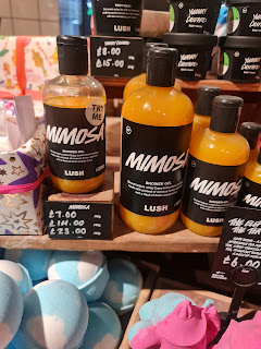 A zoomed in photo of a bunch of different sized clear bottles filled with orange liquid with black circular lids with a black label that says minosa shower gel lush in white font on a light brown rectangular shelf on a bright background