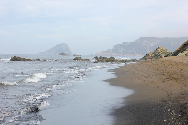 asturyjska plaża w San Juan de la Arena z widokiem na wyspę La Deva, rozległe ciemne piaski naszpikowane od czasu do czasu skałami, dzielącymi na segmenty plażę, skaliste brzegi i nagciągające od lądu du morzu mgły, nadające otoczeniu pastelowe tony