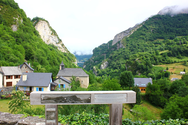 punkt widokowy na dolinę rzeki Aspe przy wjeździe do Borce, drewniana tablica informacyjna, domy o spadzistych dachach i niebisekich okiennicach z stromymi zboczami wąskiej doliny w tle