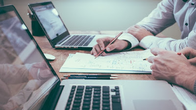 Two persons working from white notesheets with two computers and pens on the table.