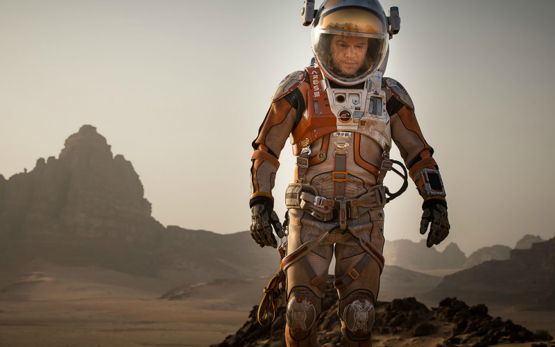 Na imagem: um astronauta caminha desolado por uma terra montanhosa e deserta, o planeta Marte. Sua roupa laranja e branca suja de terra. Ele olha para o chão.