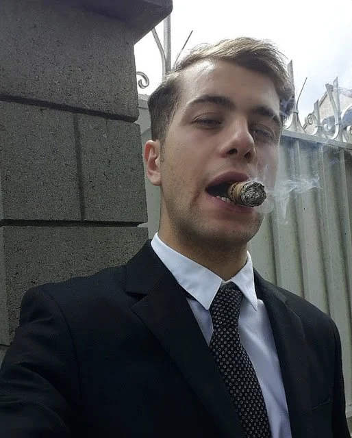 Young man with dirty blonde hair parted to the side has a cigar in his mouth wearing a suit with tie