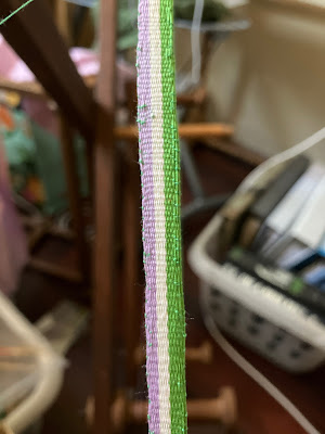a close up of a striped inkle band in progress. It is in the colours of the genderqueer pride flag (purple, white, and green) running vertically along the warp length. The weft yarn is purple. The weaving is inconsistent, with the width of the band changing three times over the four inches shown in the picture. End ID.
