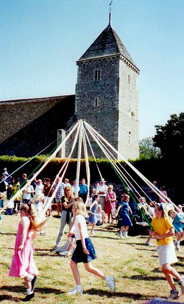 children holding ribbons run around a pole planted in the ground, in front of a church