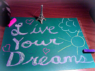 Chalk pastel on dark green paper: Live your dreams.  Two pieces of chalk pastel near the corners of the green paper.  A small Eiffle Tower figure near top of green page and a cat tail peaking in the corner of the image.  