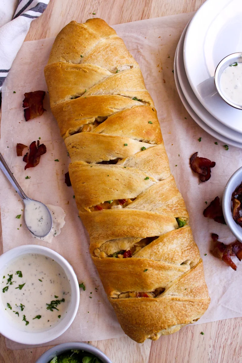 A braid of crescent rolls filled with a cheesy chicken mixture on a piece of parchment paper over a cutting board with a cup of ranch dressing sitting next to it.