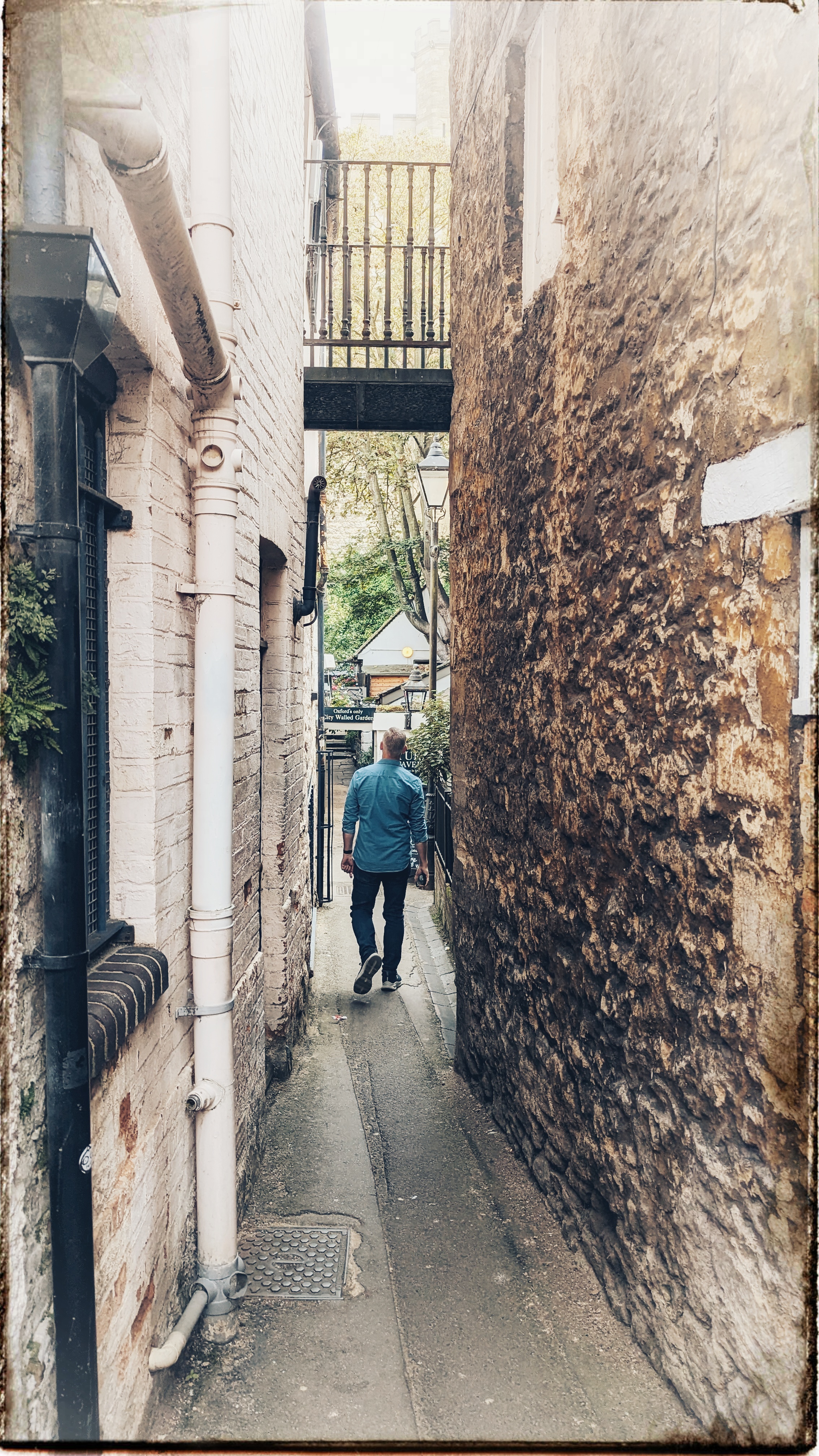 Edited and framed photo of a man in a blue shirt walking down a lane between two very old buildings. Above him a small bridge connects them.