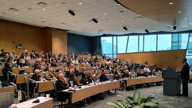 A wide view of a modern conference room filled with attendees seated at desks with microphones and laptops. The room is well-lit with natural light coming from large windows in the background. A speaker stands at the podium in the foreground addressing the audience.