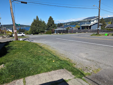 An intersection with no curbs and no sidewalks, a house with trees and  Lake Washington in the background