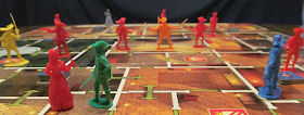 A perspective shot of the board mid-game. The board, representing various rooms in the palace of the Louvre, stretches out into the background of the photo. The four musketeer figurines, one each in blue, green, orange, and yellow, are in different rooms of the palace. The green one, in the foreground, occupies the same room as Milady D'Winter, a red figurine holding up a goblet. The yellow one, in the left midground, faces Rochefort, also in red, who holds a pistol. The blue one and the orange one are facing off against various palace guards, red figurines with swords that are scattered about the board. In the background, the grey figurine representing Queen Anne, can be seen on the far edge of the board which represents the throne room.