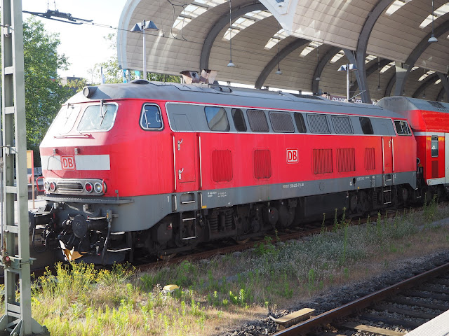 Die große rote Diesellok BR 218 steht in der Sonne vor dem Kuppeldach des Hauptbahnhof Kiel. Das Fahrwerk ist schwarz, das Chassi rot, das Dach wieder schwarz. Zwischen den Schienen wächst Gras.