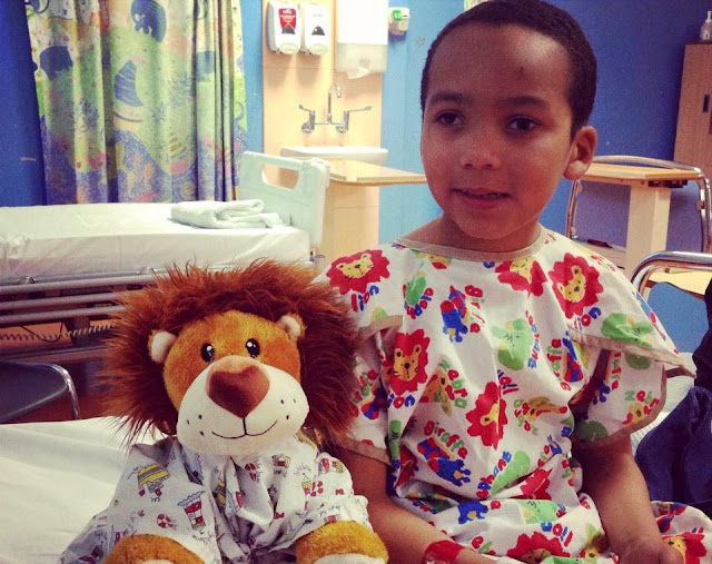 Spud in a hospital gown sat on a hospital bed with a teddy.