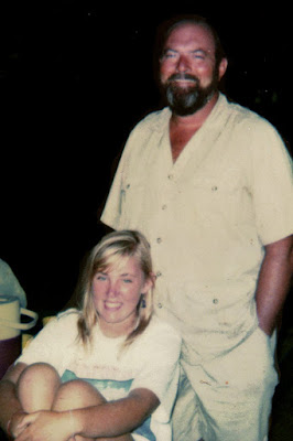 A father and daughter pose for a photo, she sits with her knees to her chest and he stands behind her both look happily at the camera.