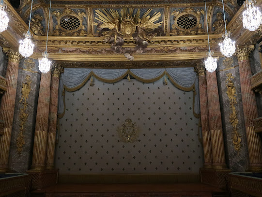 Photograph. View of a stage from the rear of a theater. The theater is extremely ornate, with carved stone and wood, lavishly painted in many colors. There are rows of crystal chandeliers to the left and right. The curtain is lowered and it's also very ornate.