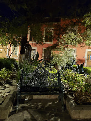 A small garden park at night light by flood lights. A wrought iron bench overlooked by a saintly statue is the focal point.