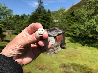 A photo of a small ceramic skull (Skulferatu 78) being held up in front of the old, tumbledown shack in Wanlockhead.  Photograph by Kevin Nosferatu for the Skulferatu Project.