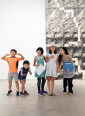 Photo of four children and an author illustrator posing in front of a large hanging art piece in a gallery