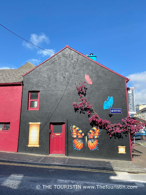 The stark black-painted facade of a corner house decorated with three large colourful butterflies and pink flowers, under a bright blue sky.