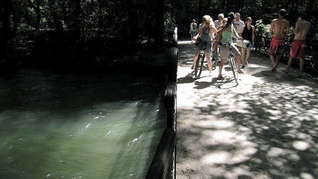 An image of a German Summer: Dark shade of the trees and cool water contrasted by bright white sunlight on a bridge with people passing by.