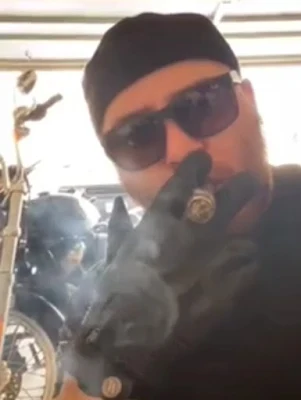 Biker wearing sunglasses and leather gloves smirking a cigar next to his motorcycle in the garage