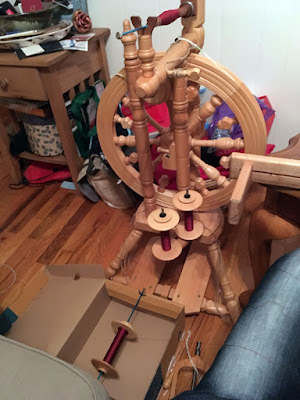 An upright wooden spinning wheel with two bobbins on the integrated lazy kate, one on a blue metal knitting needle propped in a shoebox on the floor, and a long wooden bobbin on a second blue metal knitting needle cradled by the flyer. An assortment of bags and a crowded end table fill the background; all the bobbins are thickly wound with extremely fine deep garnet thread.