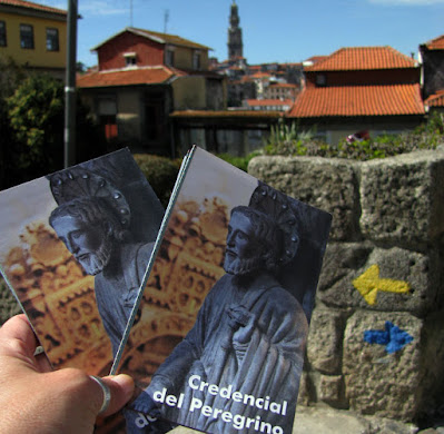 uma mão segurando duas credenciais do Peregrino de Santiago de Compostela e setas amarela e azul do Caminho de Santiago e de Fárima