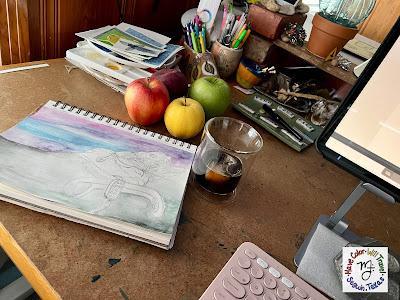 A sketchbook with a pencil and watercolor drawing of a patient in a hospital lies open on a table. It is surrounded by four colorful apples, a cup of coffee, and various art studio items.