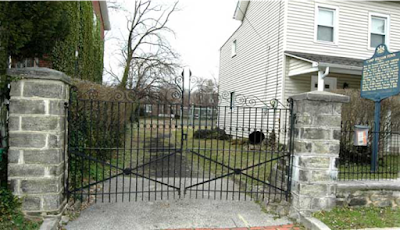 Two stone pillars with a wrought iron gate between them. A blue and gold state historical marker stands to the right.