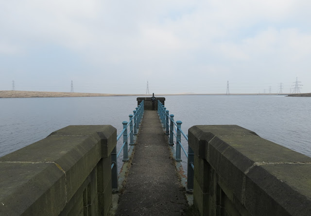 View directly down a stone jetty with blue iron railings that stretches out into the reservoir. Over the water, a line of pylons stretches across the moor.