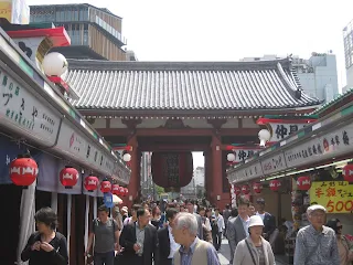 Shopping area in Asakusa. A large red paper lantern can be seen in the background of the crowd in the center of the photo housed beneath a red torii gate and green roof. On either side of the crowd are shop stalls with smaller red lanterns and paper signs written in Japanese.