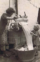 Woman using crank washer and child helping feed sheets into a wash tub