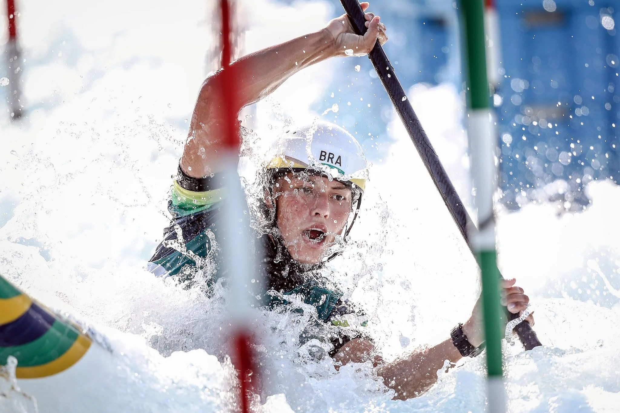 Ana Sátila competindo na canoagem slalom; ela está entre portas, com o remo na mão, de capacete, e com a água cobrindo a foto