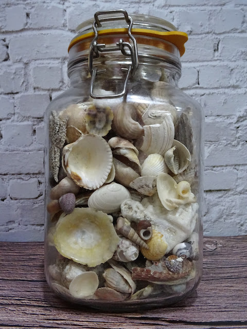 Large Kilner jar full of assorted sea shells with white brick wall behind on wooden look surface (both vinyl)