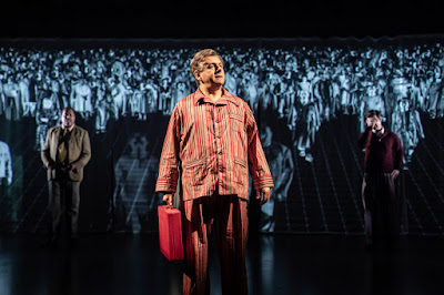 A picture taken from the play Nye showing Martin Sheen in pyjamas holding a ministerial red box with of people standing behind him