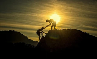 A silhouette of a boy standing on top of a hill and pullling his friend up.