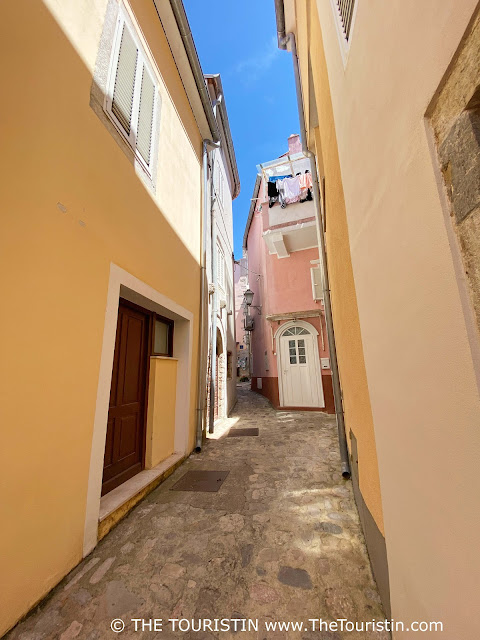 A narrow cobble-stoned lane lined by pastel-coloured houses in light yellow and pink, one with the washing in front of one window.