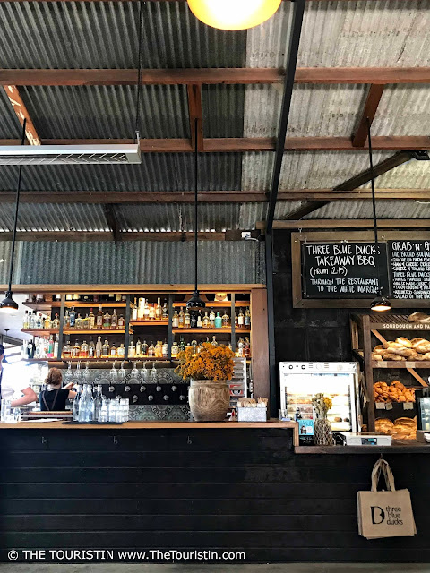 The tiled counter of a cafe under the ripple iron ceiling of a corrugated roof. Bottles and freshly baked bread on shelves in the background.