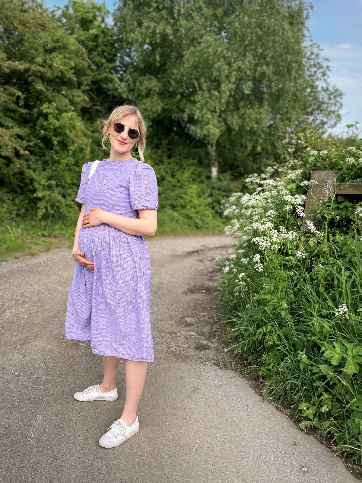 Amy is stood on a country lane in a lilac midi dress with her hands round her baby bump.