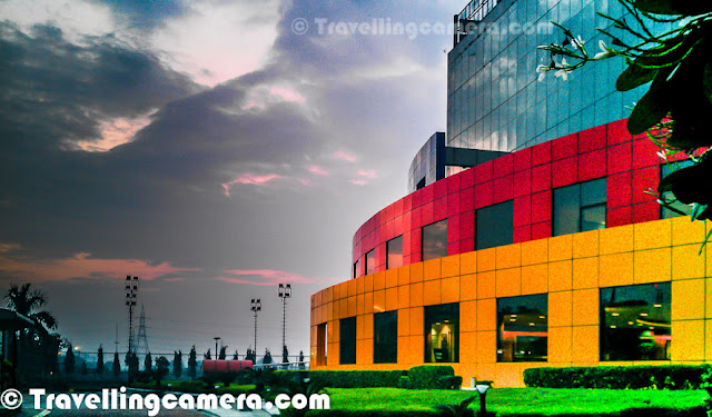Adobe India has one of the most colorful building in Noida city near to Capital city of India. This Photo Journey shares some of the mobile clicks of Adobe Noida campus from outside. Let's check out and know more about the same.Adobe Noida building with these vibrant colors was first building in sector 25A of noida. Till date, there are only two buildings in the city center sector of Noida. After one of the biggest real estate deals in Noida, Wave group has acquired rest of the sector now and building various facilities around the same. Here is a photograph of beautiful landscapes in the campus - Water fountains, green plants, colorful tiled platforms etcWhole building remains clean most of the times because of continuous cleaning efforts in campus. This building is also one of the rare green buildings in the city. Adobe Noida building has been awarded various recognitions for green efforts in the city. Adobe office in Noida is just in front of Sport-Stadium in sector 21A and Reliance building in sector 24. It's on the corner of sector 25A, facing Sector 24 on one side and sector 21A on the other side.Campus has small golf ground, half basketball court, Volleyball court & lawn tennis facilities apart from various recreational options inside the building. 