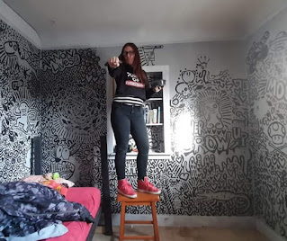 Artist Melanie Cable stands on a stool in an action pose while decorating her son's bedroom walls in a black and white graffiti-style mural.