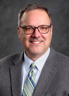 Man with glasses wears a checked shirt, striped tie and gray blazer