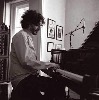 Foto em preto e branco mostra Fito Paez tocando um piano em uma pequena sala, com fotos emolduradas na parede, ao fundo.