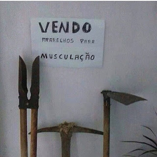 Foto: Encostado em uma parede branca, três ferramentas. Da esquerda para a direita: pá cavadeira dupla; picareta, e enxada. Acima, um cartaz branco colado na parede, escrito em letras pretas maiúsculas: Vendo, aparelhos para musculação. No canto inferior direito, parte de seis ramas finas de uma folhagem.