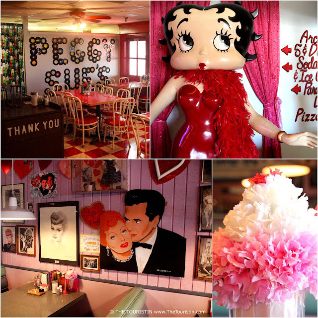 White bistro chairs around red tables on a red chequered floor in front of a wall advertising the letters Peggy Sue's. The lifesize figure of a cartoon character with a friendly round face with red lips, large blue eyes and long eyelashes, and short curly black hair dressed in a fitted red dress and a red feather boa. A pink wooden wall decorated with images of men and women and red hearts. The upper part of a pink milkshake is decorated lavishly with a pink paper flower.
