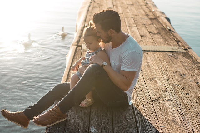 Man with young daughter sitting on a wooden jetty watching the swans swim by
