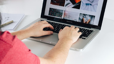 A man wearin a red shirt working his home business from a grey computer with a yellow pen on top of a white notepad by his side.