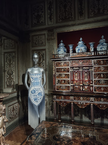 Photo of a silver mannequin in an elaborately embroidered blue and silver dress, placed in a room with a wooden floor and ornate wooden cabinetry. an inlaid stone table, and blue and white pottery.