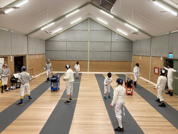 Shot of the fencing hall, with four pistes laid out with scoring boxes between, and fencers fencing on each